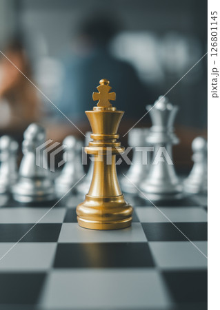 Close-up of a golden chess king on a board, surrounded by silver pieces, representing strategy and leadership in a blurred background. 126801145