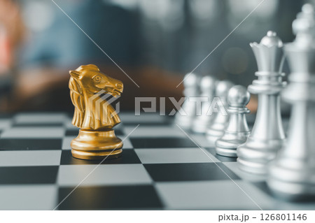 Close-up of a golden knight chess piece on a chessboard, surrounded by silver pieces, highlighting strategy and competition. Close-up of a golden knight chess piece on a chessboard, surrounded by silver pieces, highlighting strategy and competition. 126801146