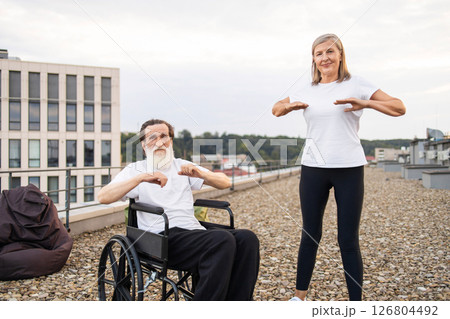 Elderly man with disability in wheelchair and senior woman stretching arms outdoors on rooftop. Health and wellness activities promoting active lifestyle for older adults. 126804492