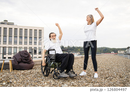 Elderly man in wheelchair and woman doing stretching exercises outdoors, promoting activity together. 126804497