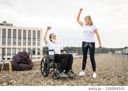 Senior couple, woman stretching, man in wheelchair raising arm outdoors on rooftop. Depicts teamwork, support, active lifestyle, disability awareness, mutual encouragement, outdoor exercise setting. Senior couple, woman stretching, man in wheelchair raising arm outdoors on rooftop. Depicts teamwork, support, active lifestyle, disability awareness, mutual encouragement, outdoor exercise setting. 126804502