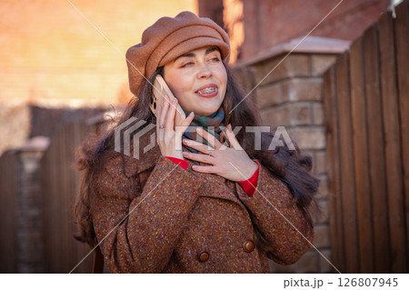 Portrait of young pretty Caucasian happy woman in a coat and hat call on phone in city street on autumn day. Lifestyle and city life 126807945