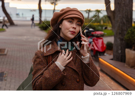 Portrait of a young beautiful Caucasian woman in a hat and coat walking down the street and talking on the phone. Concept of modern communications Portrait of a young beautiful Caucasian woman in a hat and coat walking down the street and talking on the phone. Concept of modern communications 126807969