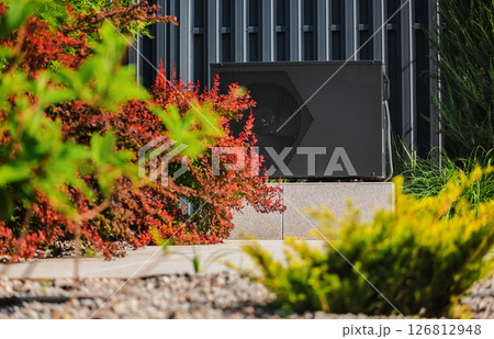 Outdoor heat pump unit placed on a concrete base in a landscaped garden, with colorful bushes and modern metal wall in the background Outdoor heat pump unit placed on a concrete base in a landscaped garden, with colorful bushes and modern metal wall in the background 126812948