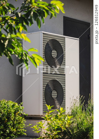 Modular heat pump unit for home heating and cooling installed next to a residential wall, surrounded by decorative plants and garden landscaping Modular heat pump unit for home heating and cooling installed next to a residential wall, surrounded by decorative plants and garden landscaping 126812949