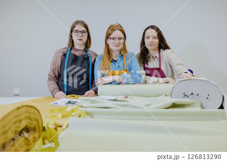 Three female seamstresses and fashion designers are standing at a table with a roll of fabric Three female seamstresses and fashion designers are standing at a table with a roll of fabric 126813290