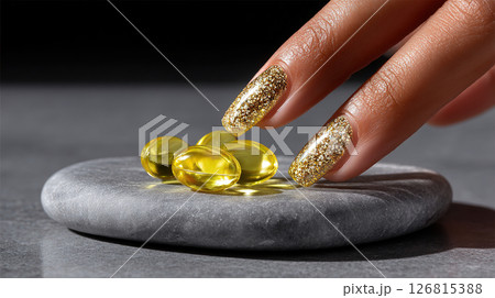 A hand with gold glitter nails gently touches omega-3 fish oil capsules on a stone. 126815388