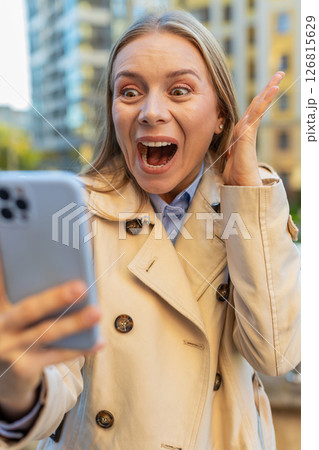 Caucasian mature woman looking shocked while reading good news on smartphone on downtown city street Caucasian mature woman looking shocked while reading good news on smartphone on downtown city street 126815629