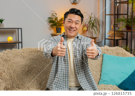 Portrait of excited smiling Chinese young man showing double thumbs up sitting on sofa couch at home 126815735