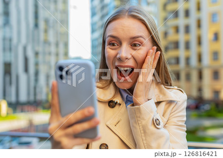 Caucasian mature woman looking shocked while reading good news on smartphone on downtown city street 126816421