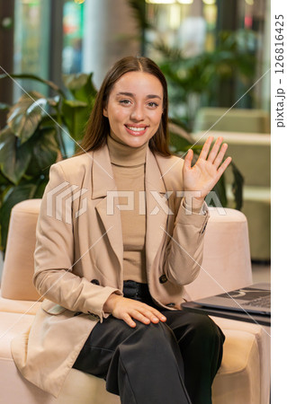 Portrait of smiling young businesswoman with laptop wave hand at camera sitting in hotel lobby 126816425