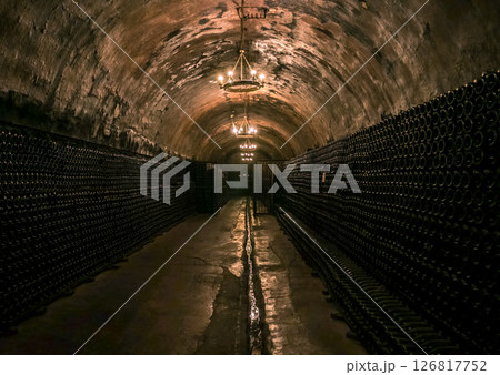 Dark underground wine cellar featuring endless rows of bottles illuminated by soft chandeliers 126817752