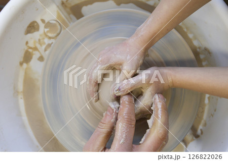 Close-up of a potter's hands and a child's hand with an item on a potter's wheel. Working with clay. Clay workshop. Craft training. 126822026