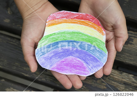 The hands of a small child are gently holding a rainbow piece of paper drawn with colored pencils. The hands of a small child are gently holding a rainbow piece of paper drawn with colored pencils. 126822046