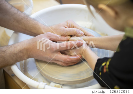 The potter teaches the craft to the child. Close-up of a potter's hands and a child's hand with a product on a potter's wheel. Working with clay. Clay master class. The potter teaches the craft to the child. Close-up of a potter's hands and a child's hand with a product on a potter's wheel. Working with clay. Clay master class. 126822052