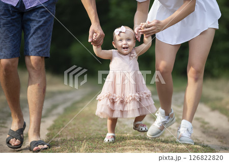 Happy one-year-old girl is being held by dad and mom. Little child walking with parents. A cute baby learns to walk with the help of her parents. 126822820