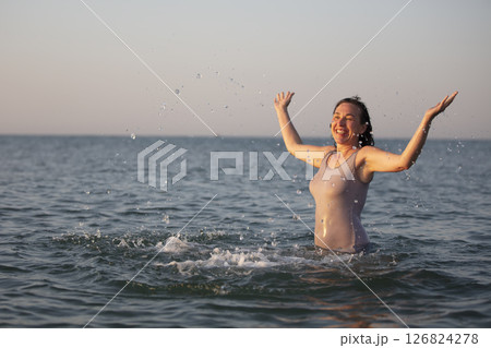 Happy woman at the sea in the sun. Woman in a swimsuit splashes water on a background of waves. Summer vacation on the beach. 126824278