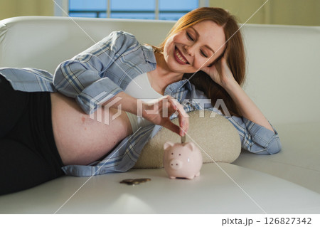 Pregnancy motherhood saving money for future investment. Pregnant woman with big belly putting coin into piggy bank at home. Pregnancy girl with piggy bank. Maternity benefit family planning concept 126827342