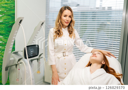 Portrait of beautician consulting with young woman client lying comfortably on modern bed, discussing innovative beauty treatments in clinic of aesthetic medicine. Concept of anti-age skincare. 126829370