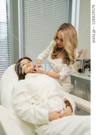Vertical portrait of blonde beautician drawing precise measurement grid on female client face, marking areas for permanent makeup procedure in contemporary salon setting. Concept of beauty treatment. 126829376