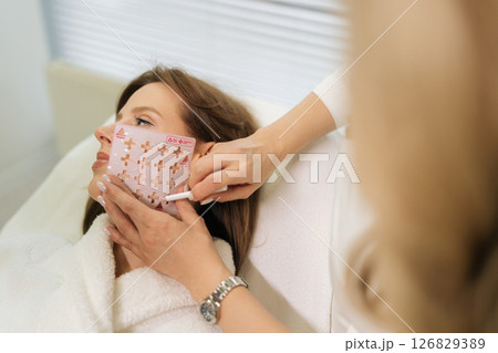 Close-up cropped shot of unrecognizable beautician using stencil to mark up woman face in preparation for anti-aging treatment in clinic of aesthetic medicine. Concept of non-surgery cosmetology. 126829389