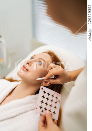 Close-up of beautician marking facial injection points with white pencil and transparent grid, prepping patient for cosmetic treatment in clinic of aesthetic medicine. Concept of anti-age skincare. 126829393
