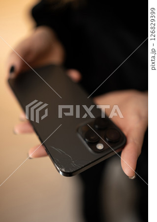 Vertical cropped shot of unrecognizable woman holding broken smartphone with cracked back glass, checking damaged surface with finger. Sad female touching cracked damaged back glass of phone. Vertical cropped shot of unrecognizable woman holding broken smartphone with cracked back glass, checking damaged surface with finger. Sad female touching cracked damaged back glass of phone. 126829399
