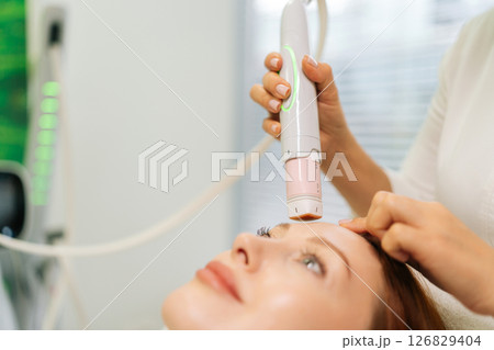 Close-up cropped shot of dermatologist using modern device performing mesotherapy procedure on forehead of woman lying on treatment table in beauty salon. Concept of anti-age rejuvenation skincare Close-up cropped shot of dermatologist using modern device performing mesotherapy procedure on forehead of woman lying on treatment table in beauty salon. Concept of anti-age rejuvenation skincare 126829404