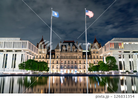 Night view of the New York State Capitol in Albany, framed by modern government buildings and flagpoles, with dramatic reflections on the still water of Empire State Plaza under a cloudy sky. Night view of the New York State Capitol in Albany, framed by modern government buildings and flagpoles, with dramatic reflections on the still water of Empire State Plaza under a cloudy sky. 126831354