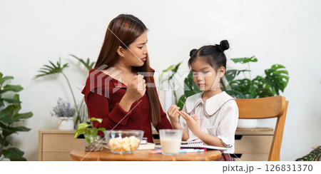 Engaging Learning Moments. A mother patiently guides her daughter through school preparations over snacks. Engaging Learning Moments. A mother patiently guides her daughter through school preparations over snacks. 126831370