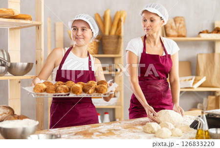 Two professional baker working in bakehouse. Happy adult woman kneading dough and young girl arranging freshly baked bakery products Two professional baker working in bakehouse. Happy adult woman kneading dough and young girl arranging freshly baked bakery products 126833220