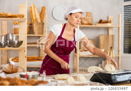 Middle-aged female baker cutting and weighing dough on scales in bakehouse Middle-aged female baker cutting and weighing dough on scales in bakehouse 126833497