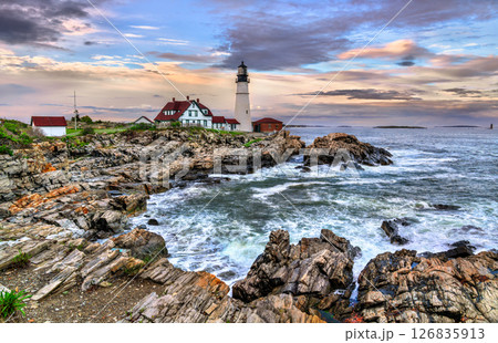 Portland Head Light stands prominently on the rocky shore of Cape Elizabeth, Maine, USA framed by dramatic ocean waves and colorful evening skies. The historic lighthouse and its red-roofed buildings 126835913