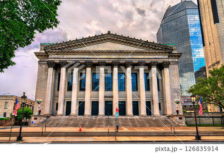 Symphony Hall in Springfield stands with a stately colonnaded facade in Greek Revival style, flanked by American flags and surrounded by a mix of historical and modern architecture. The dramatic cloud 126835914