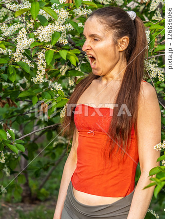 Young woman sneezes in the park against the background of a flowering tree. Allergy to pollen concept. Young woman sneezes in the park against the background of a flowering tree. Allergy to pollen concept. 126836066