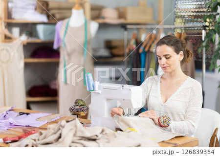 Working in sewing workshop - female dressmaker working at sewing machine making stitches on fabric 126839384
