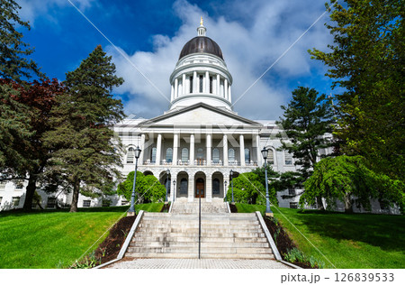 Classical white granite capitol building with a prominent dome and columned portico, surrounded by manicured green lawns and tall trees in Augusta, Maine, USA Classical white granite capitol building with a prominent dome and columned portico, surrounded by manicured green lawns and tall trees in Augusta, Maine, USA 126839533