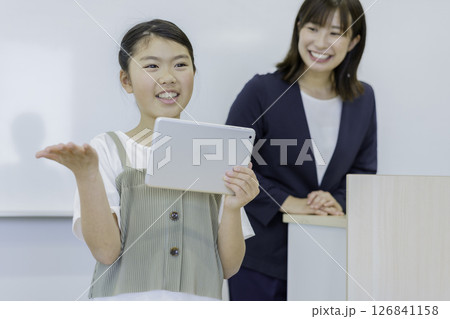 タブレットを持って発表する小学生の女の子と先生　撮影協力：学校法人電波学園 東京電子専門学校 126841158