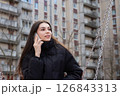 Beautiful Caucasian young woman wearing a black down jacket talking on smartphone while sitting on a swing near urban buildings 126843313