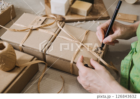 Person writes on a kraft tag attached to a ribbon-wrapped gift box near handmade soap. Handwritten tags, small business packaging, emotional branding, customer connection 126843632