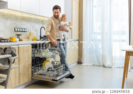 Young father putting plates into the washing machine while holding his son Young father putting plates into the washing machine while holding his son 126845658