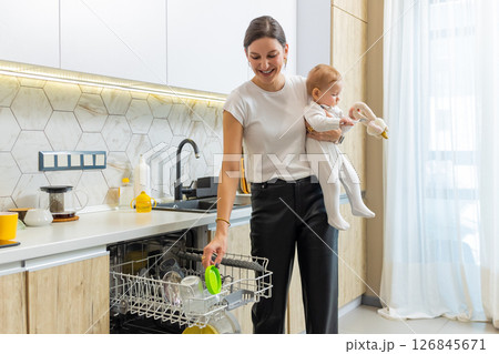 Young mother putting plates into the washing machine while holding his son Young mother putting plates into the washing machine while holding his son 126845671
