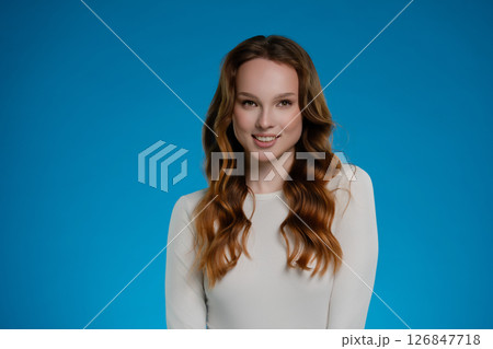 Woman with long hair poses in studio against a vibrant blue background, expressing a warm and inviting emotion Woman with long hair poses in studio against a vibrant blue background, expressing a warm and inviting emotion 126847718