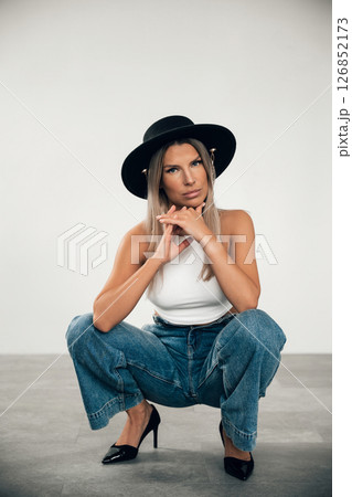 Fashion model in stylish outfit squatting indoors with a serious expression and bold hat 126852173