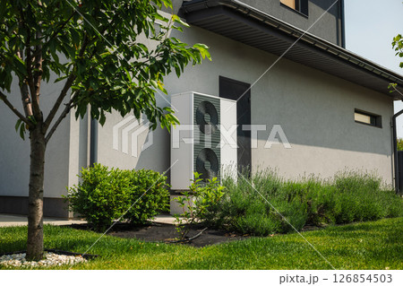Modern heat pump unit installed next to a residential house wall, surrounded by green lawn, bushes, and young trees in a landscaped yard Modern heat pump unit installed next to a residential house wall, surrounded by green lawn, bushes, and young trees in a landscaped yard 126854503
