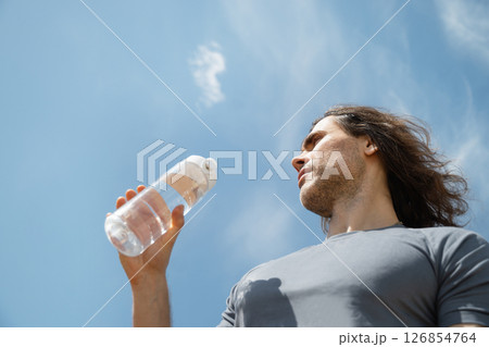 Handsome curly brunette man wear gray t-shirt with water bottle against blue sky background. Summer heat weather. Outdoors. Hero view. Close up. Banner. 126854764