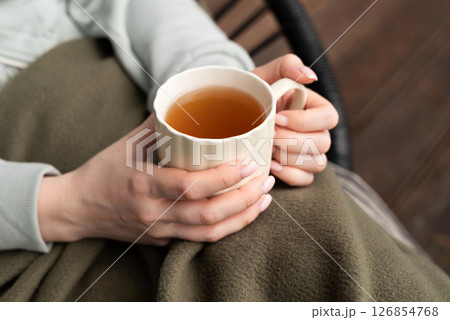 Woman holding a cup of hot tea while sitting cozy under a blanket, warming hands on a chilly day. Woman holding a cup of hot tea while sitting cozy under a blanket, warming hands on a chilly day. 126854768