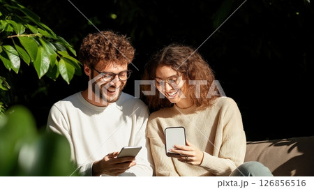 Carefree Young Couple Relaxing and Enjoying Time Together Outdoors in a Lush Green Park or Garden Setting While Using Their Smartphones 126856516