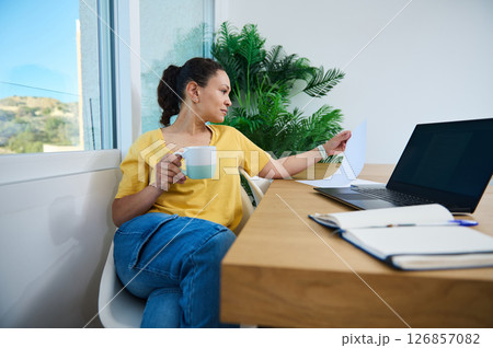 Businesswoman Reviewing Documents While Enjoying Coffee in Home Office Businesswoman Reviewing Documents While Enjoying Coffee in Home Office 126857082