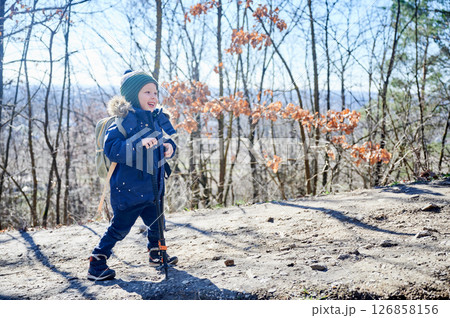 Young child smiling while hiking on forest trail, bundled up in blue winter jacket with fur-lined hood and green striped beanie. Kid holding two trekking poles and enjoying sunny day outdoors. Young child smiling while hiking on forest trail, bundled up in blue winter jacket with fur-lined hood and green striped beanie. Kid holding two trekking poles and enjoying sunny day outdoors. 126858156